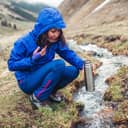 A woman dressed in weatherproof clothing filling a metal bottle with water from a stream outdoors