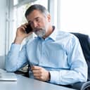 A middle-aged businessman sitting in chair looking at laptop having a conversation on smartphone
