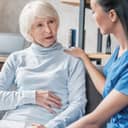Female nurse in uniform attentively listening to elderly patient's stomach pain complaints at home