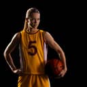 A young woman in a basketball uniform, holding the ball in one hand and standing against a black background.