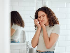 A young woman in a bathroom holding her cheek because of a toothache 