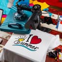 top view of a workspace with equipment for heat pressing designs onto shirts with other tools around and a T-shirt with 'I love summer' print on it