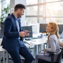 young businessman lounging on a desk and talking with a female colleague who is seated at her desk in the office