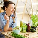 Young and happy woman eating healthy salad sitting on the table with green fresh ingredients indoors