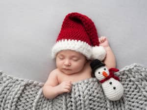 A baby sleeping covered with a blanket wearing a wool Santa Clause cap and a snowman toy