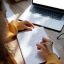 A young woman writing in her notepad with a laptop in front of her