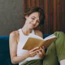 Portrait of a cheerful woman in a white tank top relaxing with a book on a blue couch.