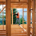 male carpenter hammering nails while wearing a helmet and vest in a two-story wooden house near a forest