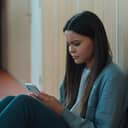Sad teenage girl reading a text message on her phone while sitting on the floor in a hallway.