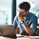 A tired young man rubbing his eyes while sitting in front of his laptop at an office