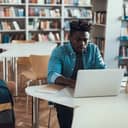 A focused student is sitting and studying in the library, with a laptop in front of him.