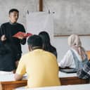 High school students attentively listening as a young male teacher explains something at the front of the classroom.
