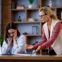an angry female boss scolding a young female employee who is seated at her desk in the office