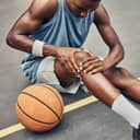 A basketball player sitting on the court next to a basketball holding his knee