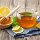 A glass cup of tea with lemon slices, mint and a honey in a bowl placed on a cloth on a wooden table