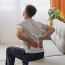 Man sitting on a couch at home in the morning, reaching for his lower back and exhibiting discomfort from a muscular strain.