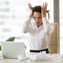 Panicked woman at work looking at her laptop with lots of crumpled paper lying around on the desk