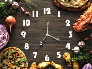 A time to eat concept with a clock on a wooden surface with food surrounding it