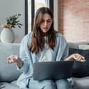 An annoyed young woman sitting on a couch in her living room, visibly frustrated while working on her laptop.