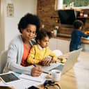 Woman doing work on kitchen table while a child is in her lap while another child is by the kitchen work surface