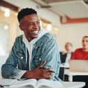 A male university student sitting in a classroom, gazing away from his desk with a smile.