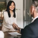 Business, career and placement concept - image from back of two employers sitting in office and shaking hand of young asian woman after successful negotiations or interview