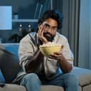 bored man with a beard and glasses watching TV at home, eating popcorn and sitting in his living room