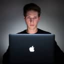 Man sitting at a Macbook computer in a dark room.