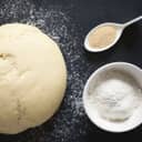 Rolled up dough, spoonful of yeast and bowlful of flour on black surface