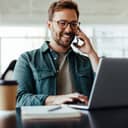 A smiling man with sunglasses talking on the phone while working on his laptop