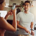 Silly, goofy and interracial couple taking pictures, having fun and cooking together in the kitchen at home. Happy girlfriend taking pictures of boyfriend, making funny faces and preparing dinner