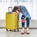 A tired woman leaning on her head on a yellow suitcase holding a red passport sleeve