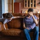 Small boy leaning over sofa hand rest reaching for a man's shoulder while tries to cover his face with left hand