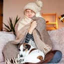 woman wrapped in a blanket and winter attire sitting on a couch in her living room, with her dog lying in her lap