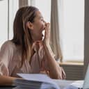 Woman sitting in front of a laptop at desk yawning with her hand over the mouth and looking through a window