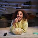 A woman smiling at the camera lying on the living room carpet eating popcorn.