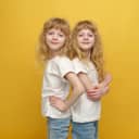 Portrait of blonde twin girls, standing back-to-back against a yellow backdrop.
