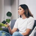 A confused woman holding a remote while sitting on a sofa