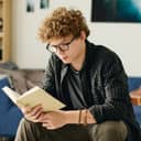 close-up of a male teen reading a novel while seated on a bed in his bedroom