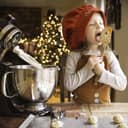 A little girl in the kitchen making holiday cookies, licking the dough from a spatula.