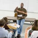 A teacher leaning on a desk giving a lecture to high-school students