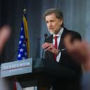 older politician speaking at a press conference with American flags in the background