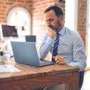 middle-aged businessman sitting at the office with his laptop in front of him, looking worried about something he's reading on the screen