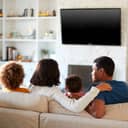 Close-up back view of a young family of four cozily seated on a couch, watching TV in their living room.