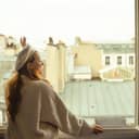 beautiful woman dressed in a beige sweater and beret standing by a window and waving to someone, Parisian rooftops in the background