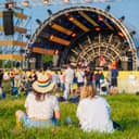 The back view of two friends sitting on the grass at an open-air music festival concert.