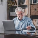 An older man sitting at desk behind a laptop taking down notes on a pad