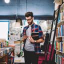 Stylish young man in reading a book's synopsis on his mobile phone while spending time in a bookstore.