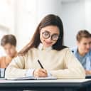 Portrait of a happy girl wearing glasses, sitting at a table in a high school, focused on writing notes in her notebook.