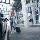 Beautiful young woman standing with a trolley suitcase next to the car at airport terminal.
